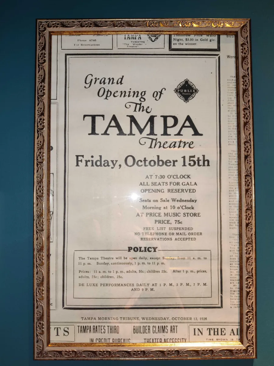 Framed newspaper clipping announcing the grand opening of the Tampa Theatre on Friday, October 15th at 7:30 PM, from the Tampa Morning Tribune dated Wednesday, October 13, 1926. The advertisement includes details about seat reservations, ticket prices (75 cents), and theater policies. The ornate wooden frame has decorative carved borders.