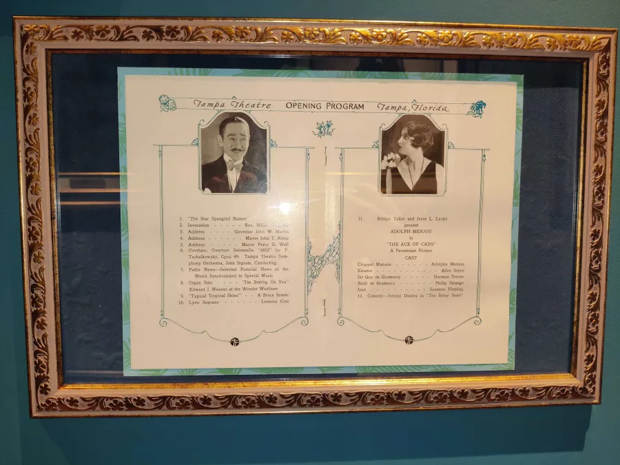 Framed Tampa Theatre opening night program, featuring two sepia-toned portrait photographs of performers flanking a detailed program listing.  The left portrait shows a man with a mustache in formal attire, while the right shows a woman with dark hair. Text lists various musical numbers, performances, and cast members for 